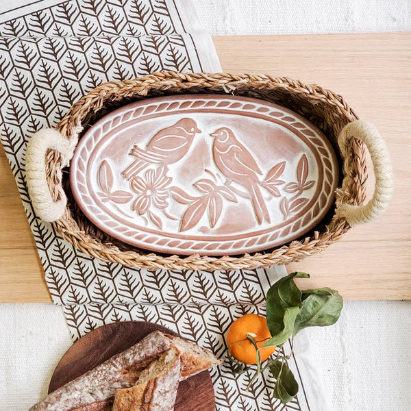 Decorative terracotta bread warming tray with lovebirds design in a wicker basket on a wooden surface.