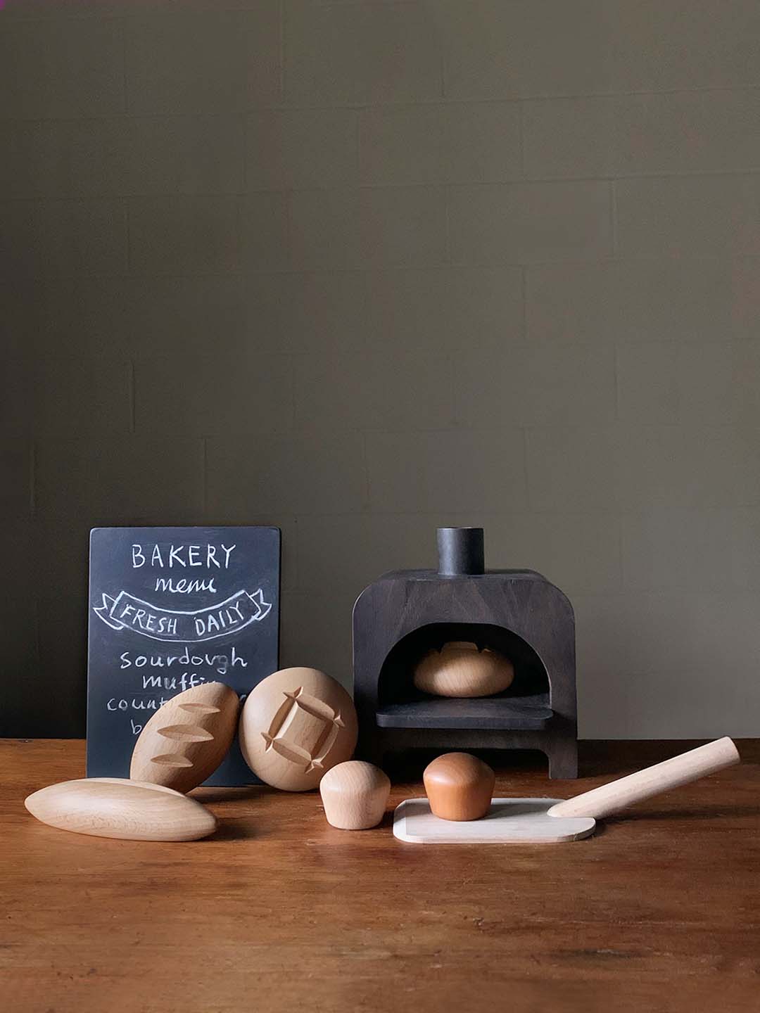 Children's wooden play bakery set with a small oven, bread, spatula and chalkboard sign.